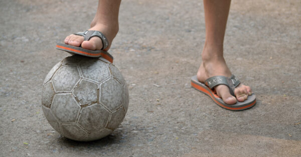 pés de um garoto com chinelo pisando na bola de futebol